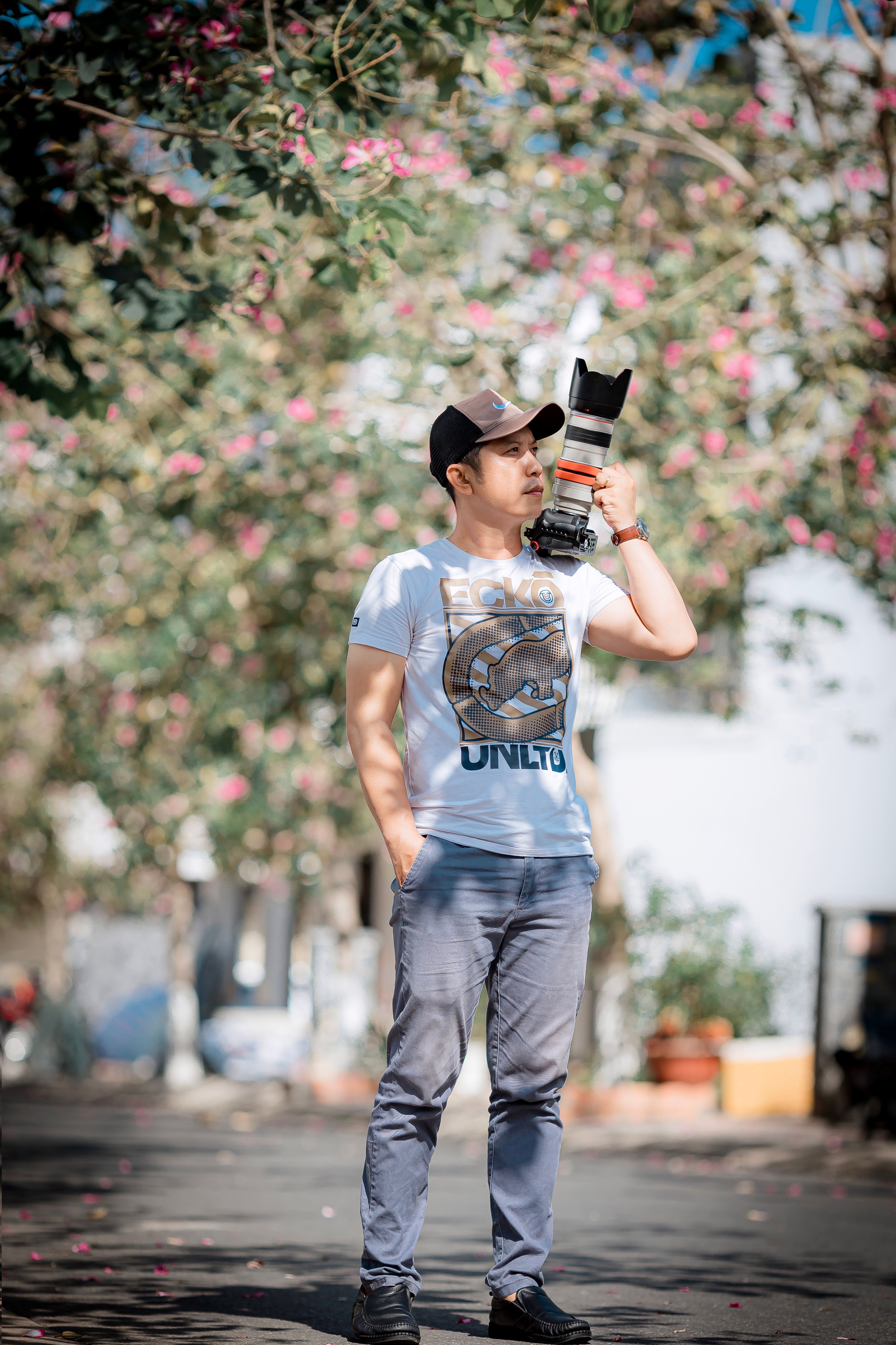 Man with a camera standing on a flower-lined street under sunlight.