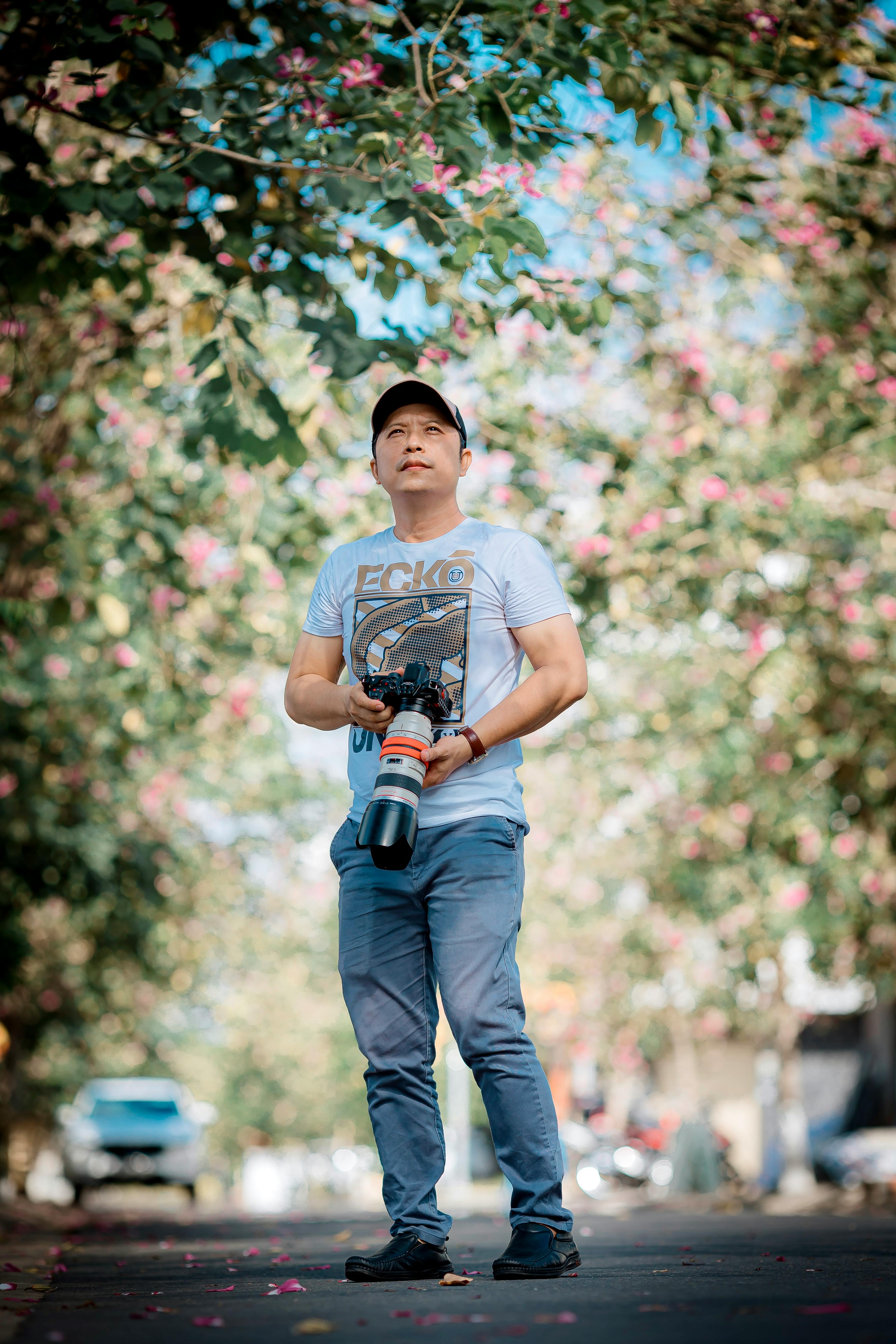 Man with camera stands under flowering trees on a sunny day.