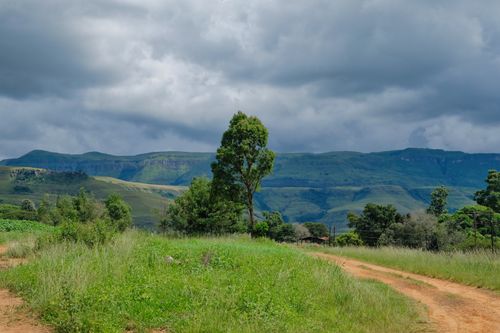 Free A picturesque dirt road leading through lush green hills and mountains under a cloudy sky. Stock Photo
