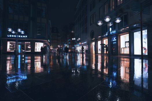 Moody urban night shot with long reflections on wet city streets, illuminated storefronts, and silhouetted pedestrians.
