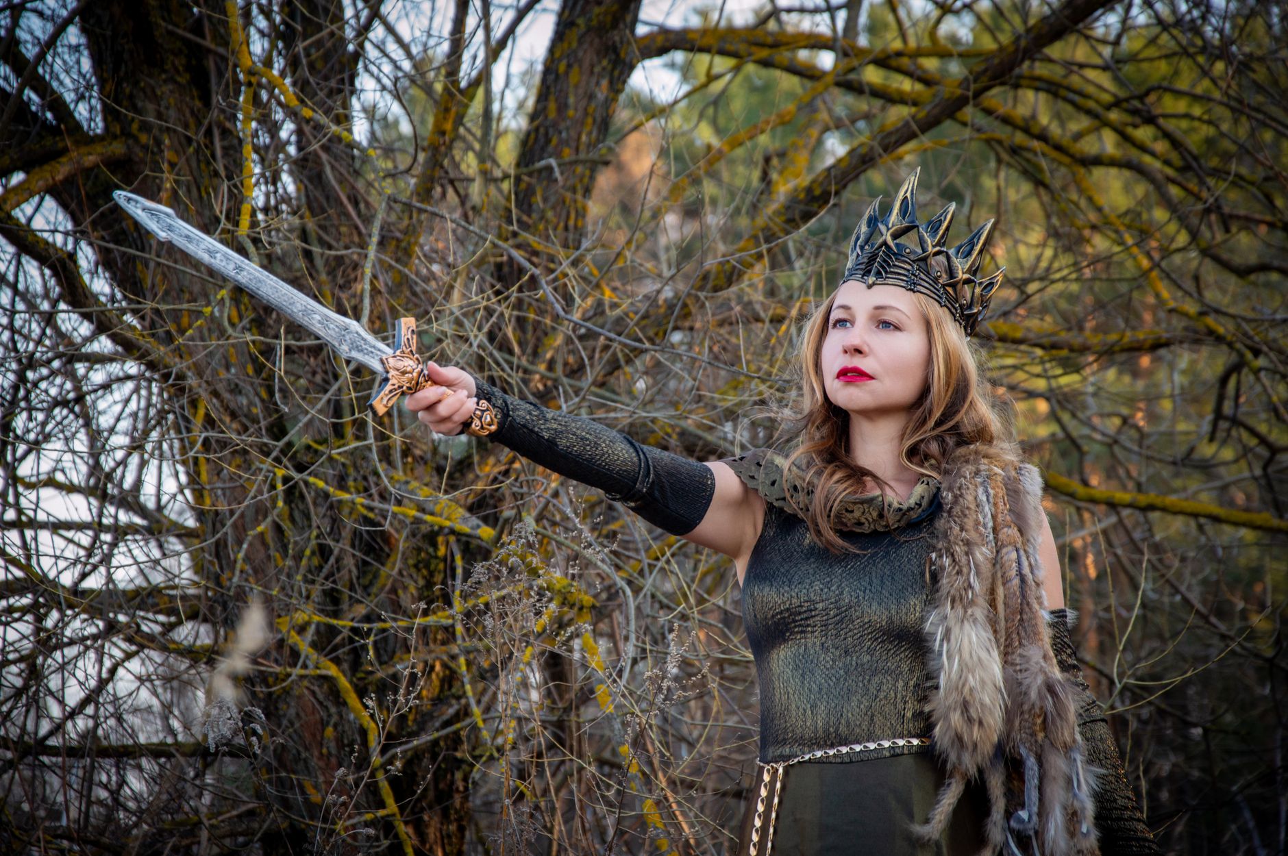 A fierce warrior queen in medieval costume holding a sword against a forest backdrop.