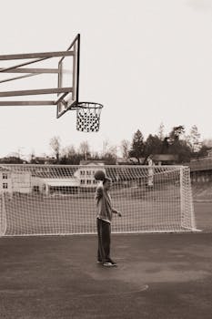 Black and white photo of a teenager playing basketball on an outdoor court with an empty net in the backdrop.
