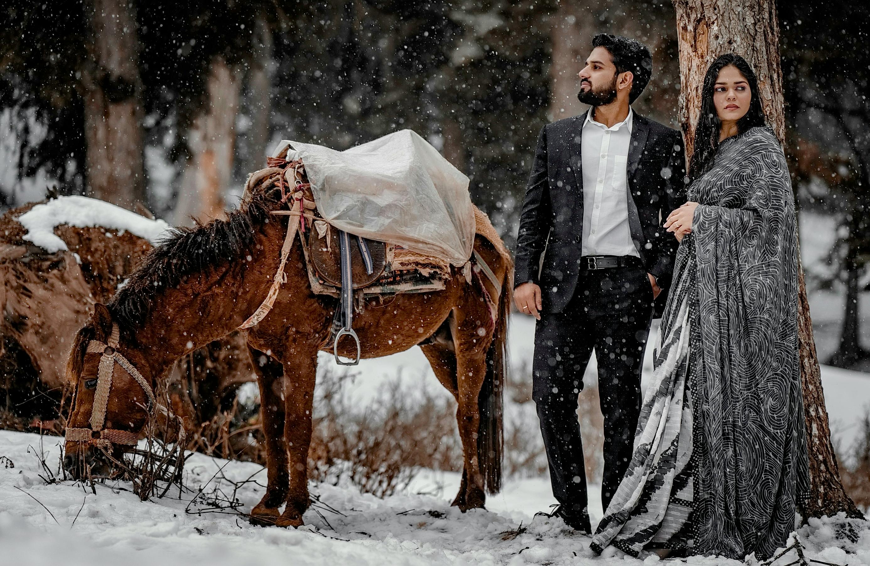Elegant couple in formal attire standing with a horse in a snowy forest setting.