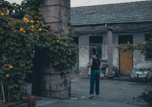A woman stands quietly in a rustic courtyard covered with lush vines and yellow flowers.