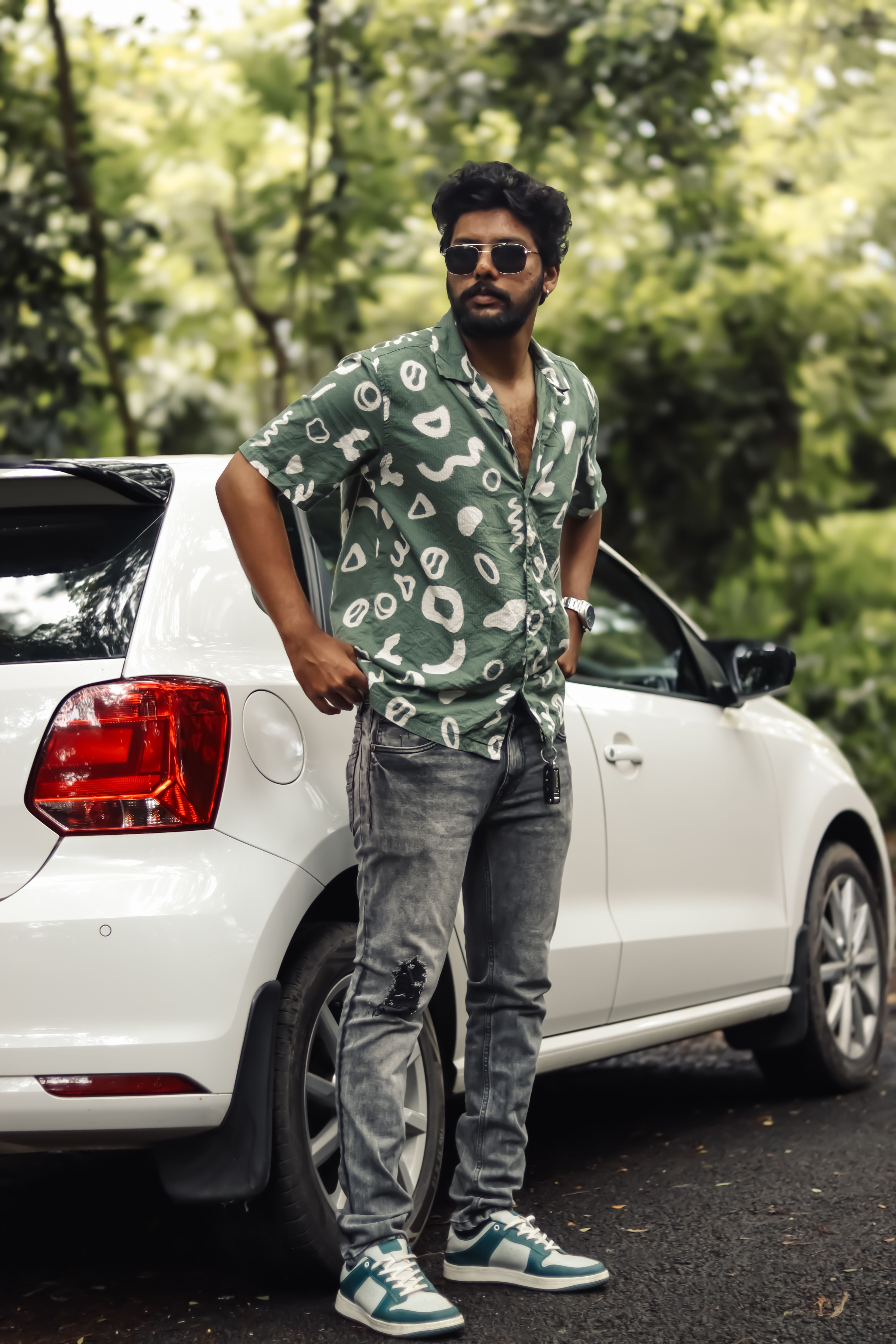 Stylish man in a green patterned shirt poses by a car in a lush, urban environment.