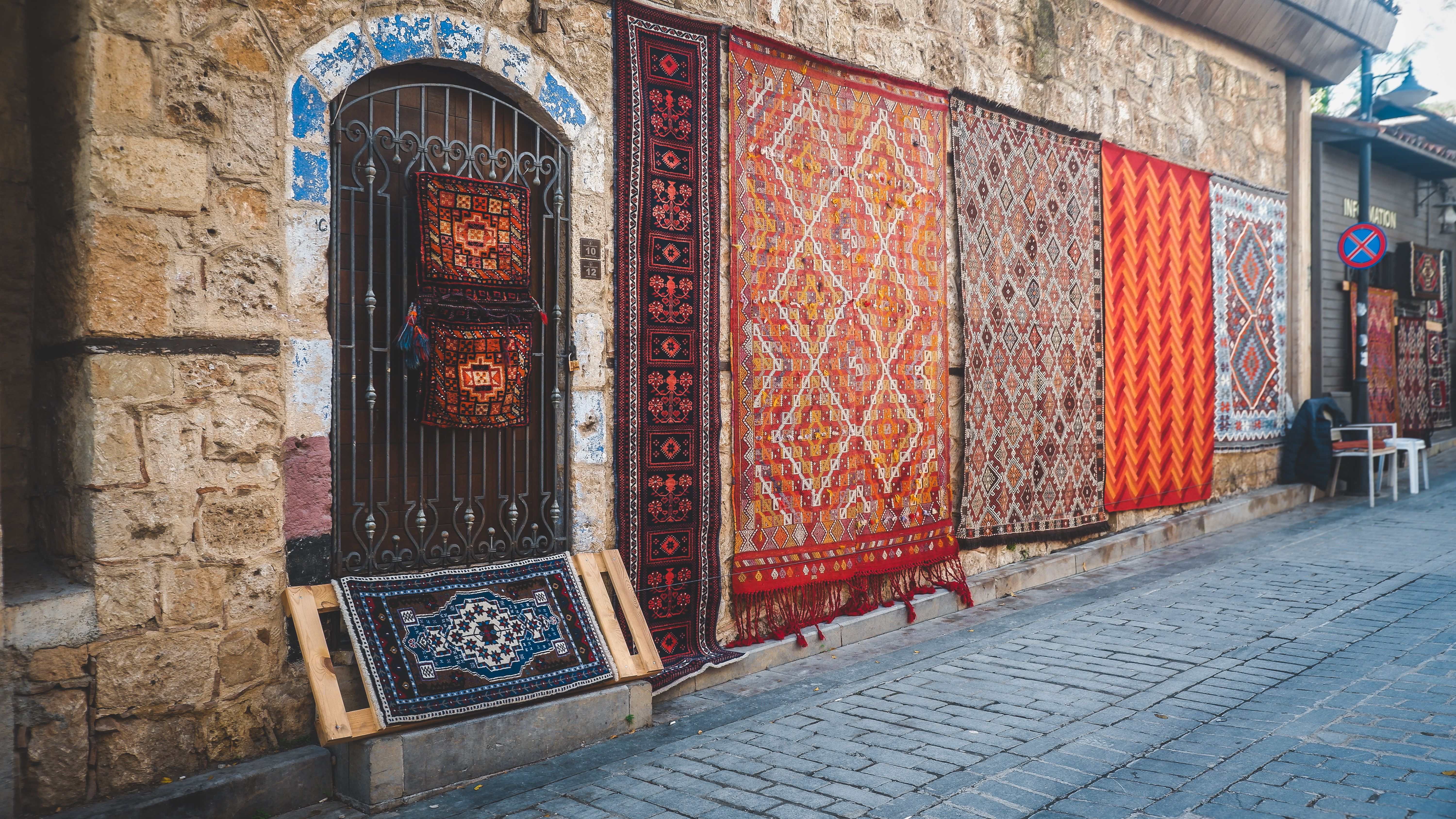 Vibrant traditional rugs hanging on a rustic stone wall in an outdoor market setting.