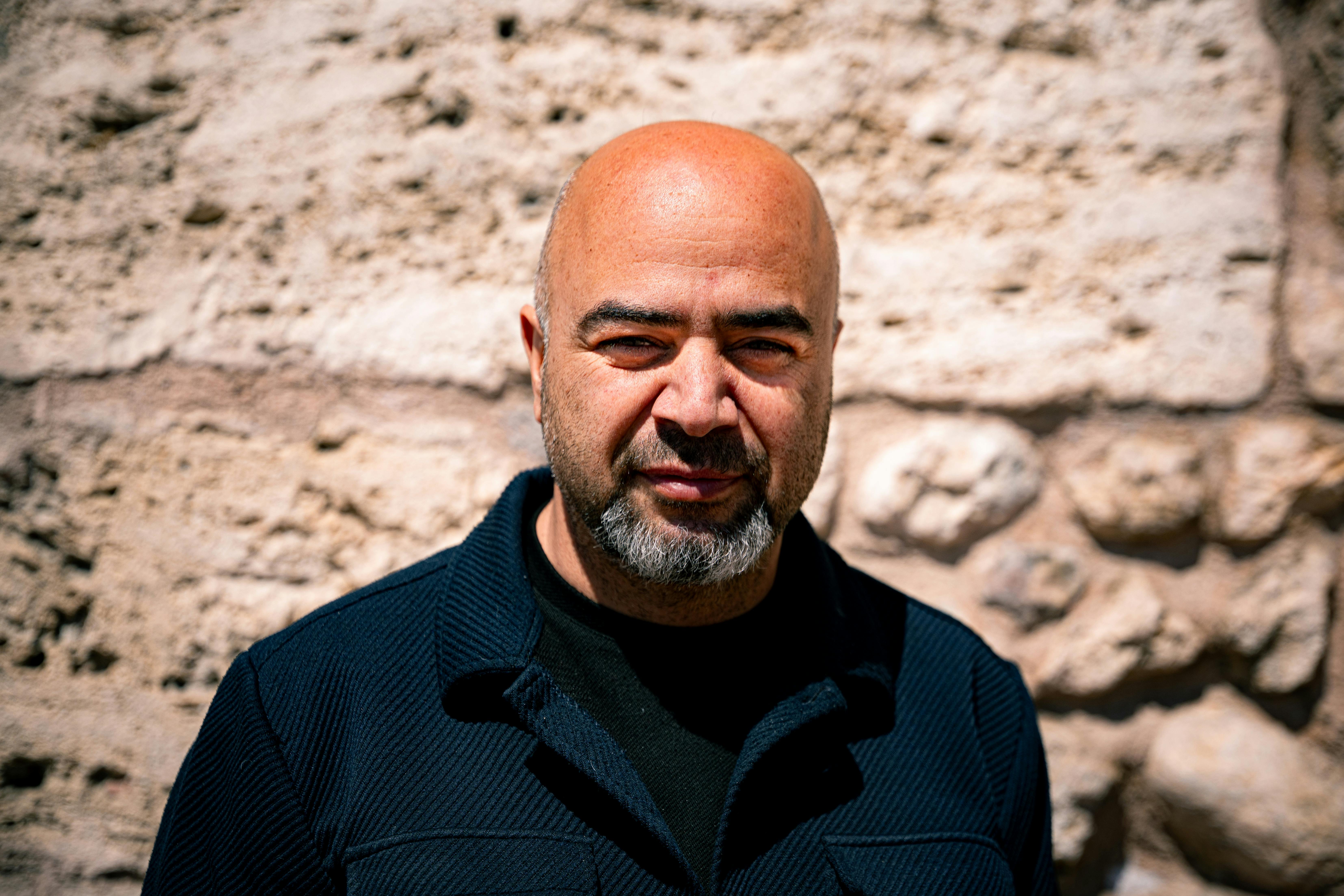 Close-up portrait of a bald man with a beard standing outdoors against a rustic stone wall.