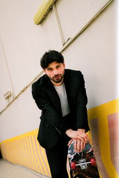 Stylish man in black suit leaning with skateboard outdoors against a wall.