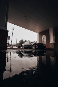 A black car parked under a building canopy with its reflection visible in a puddle on a rainy day.