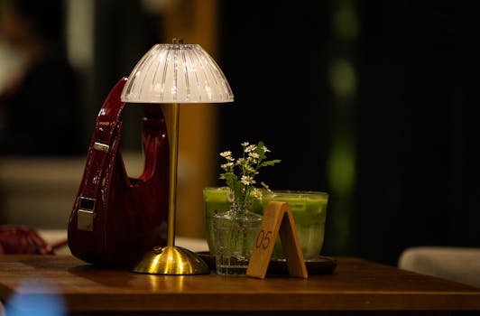 A warm cafe table setting with a lamp, handbag, green beverage, and flowers.