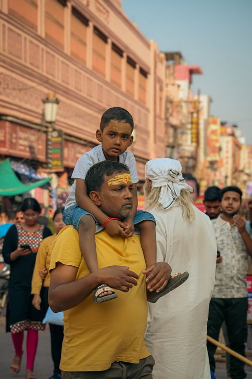 Free A father carries his son on shoulders in a bustling Varanasi street market, showcasing local culture. Stock Photo