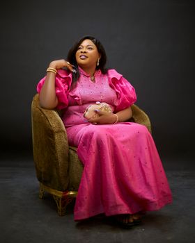 Sophisticated woman in pink attire sitting on an armchair in a studio setting.