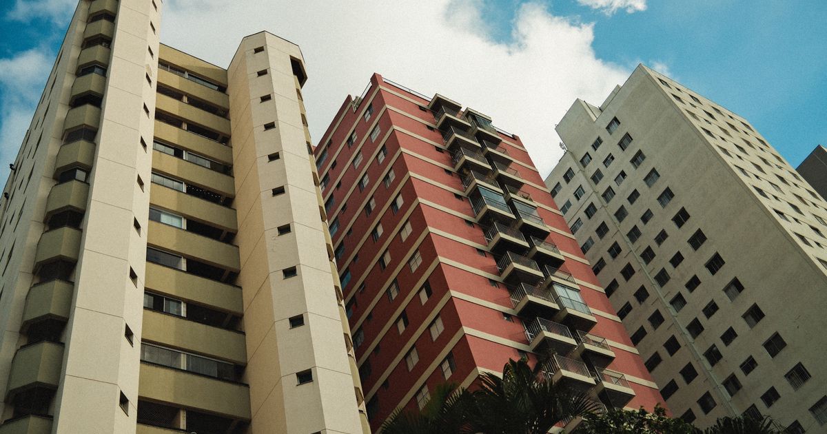 Uma vista em ângulo baixo de três prédios altos com arquitetura diversificada sob um céu azul claro, destacando a vida urbana.