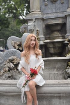 Young woman in a white dress holds red roses while sitting by an ornate fountain.