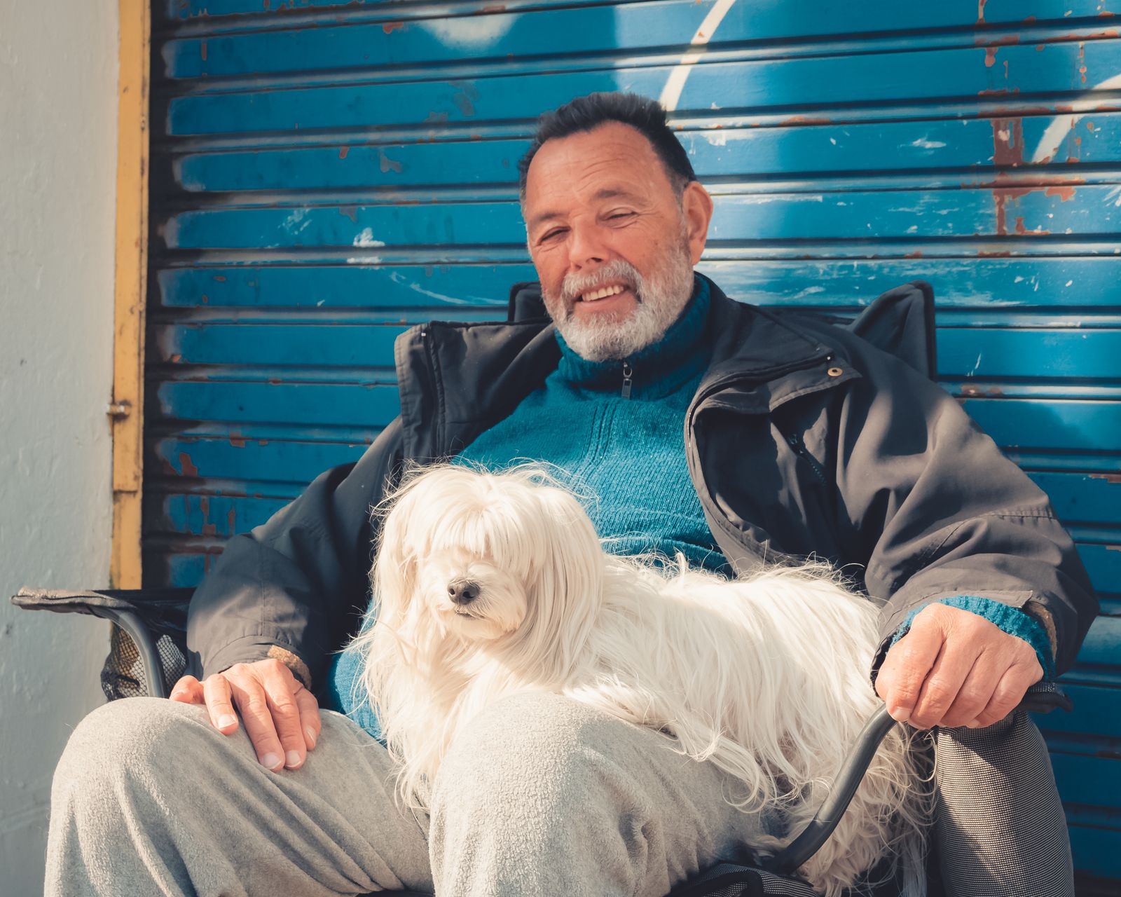 A smiling grey-bearded man sitting with a fluffy white dog on his lap in front of a blue shutter
