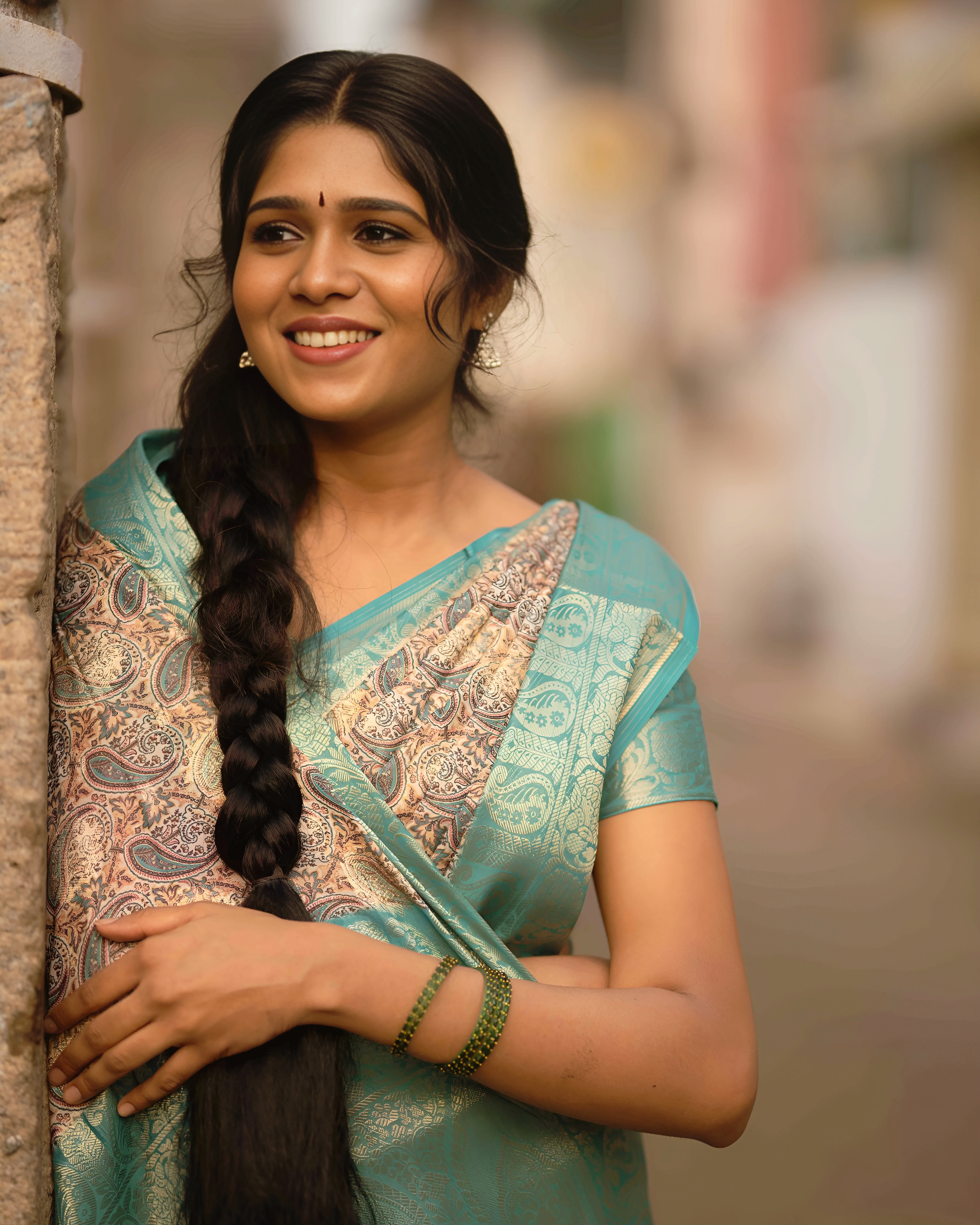 Woman in a green saree smiling while leaning on a wall outdoors.