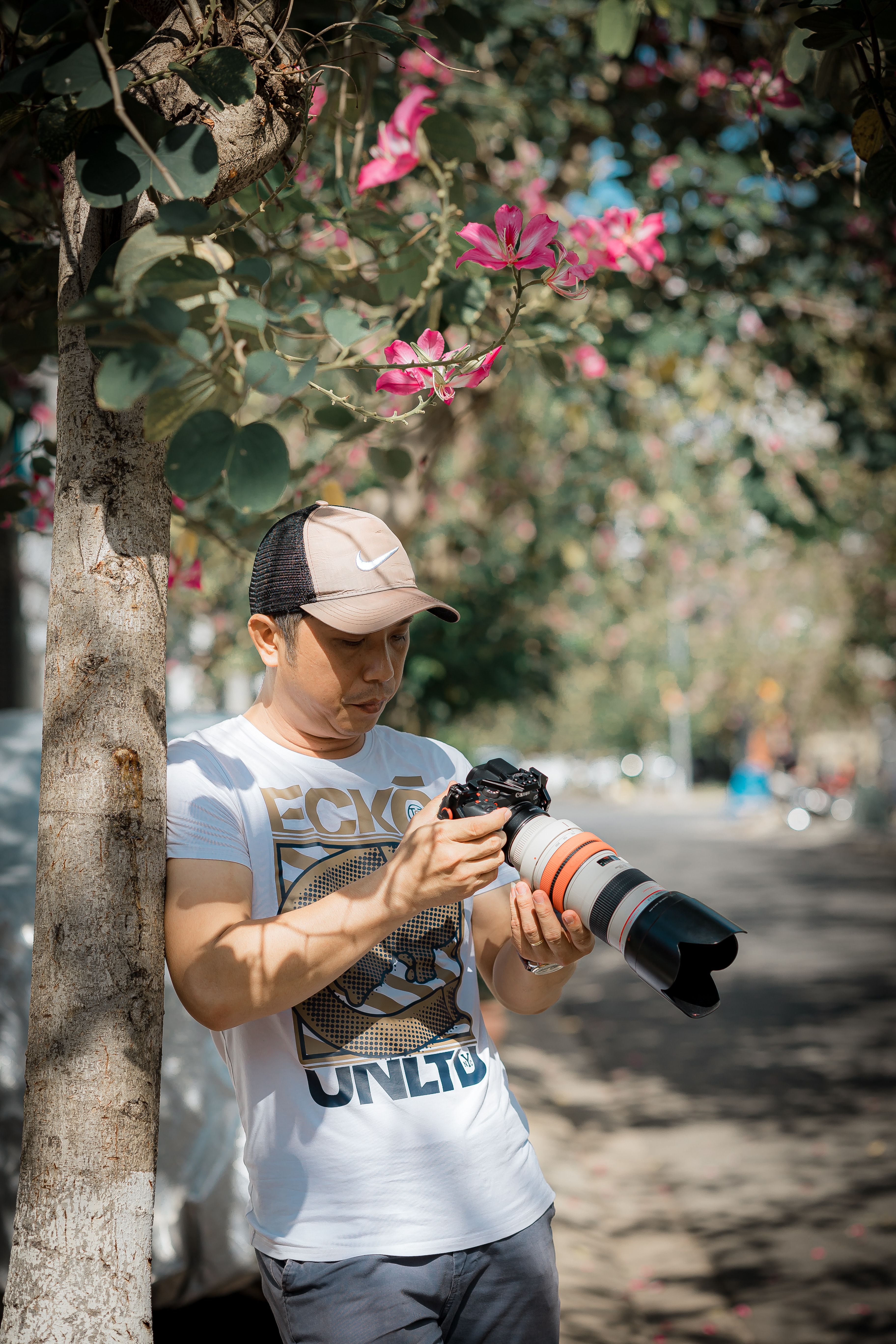 Man leaning against tree with camera in blooming urban setting.