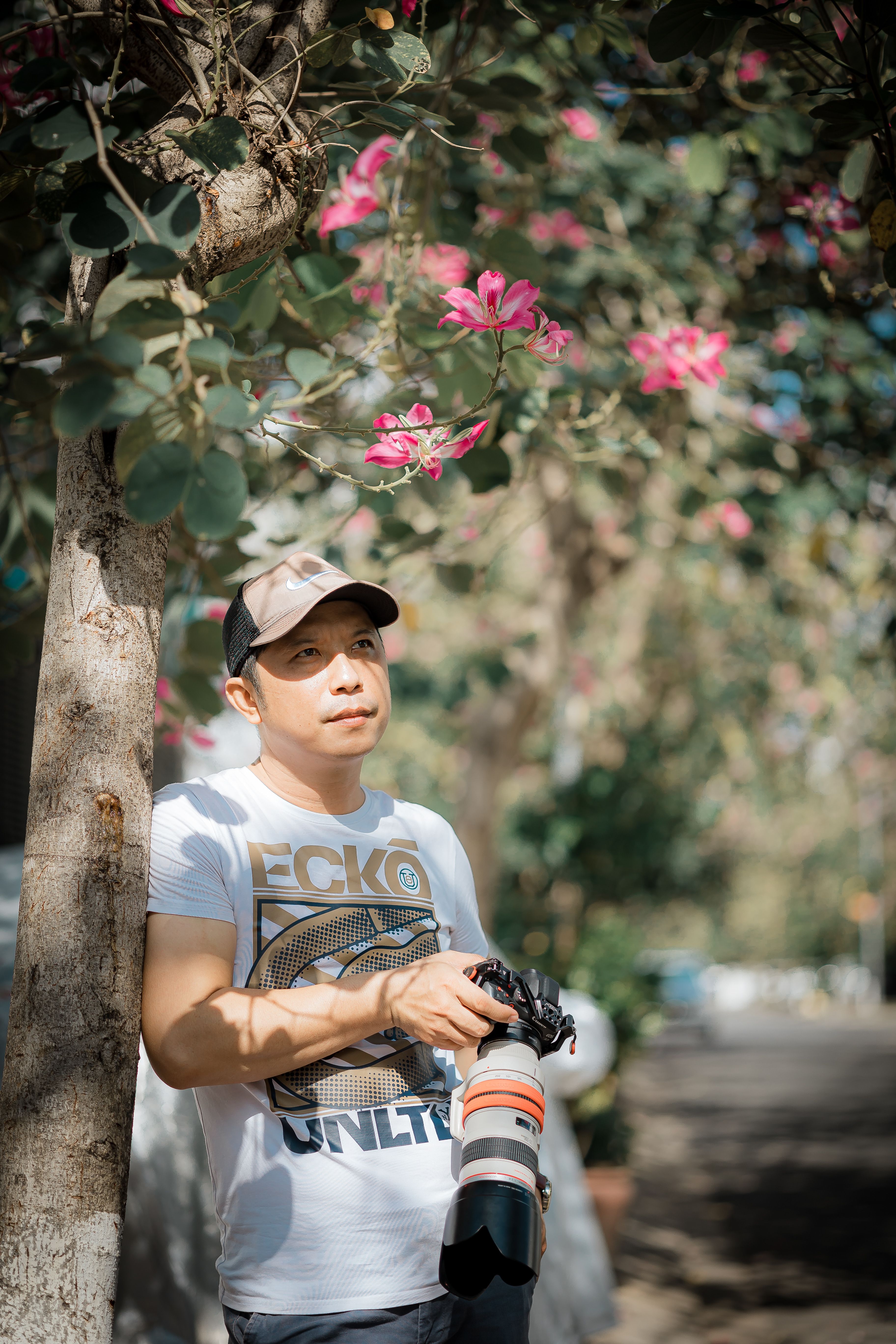 A photographer holds a camera under a blooming tree, enjoying outdoor sunlight.