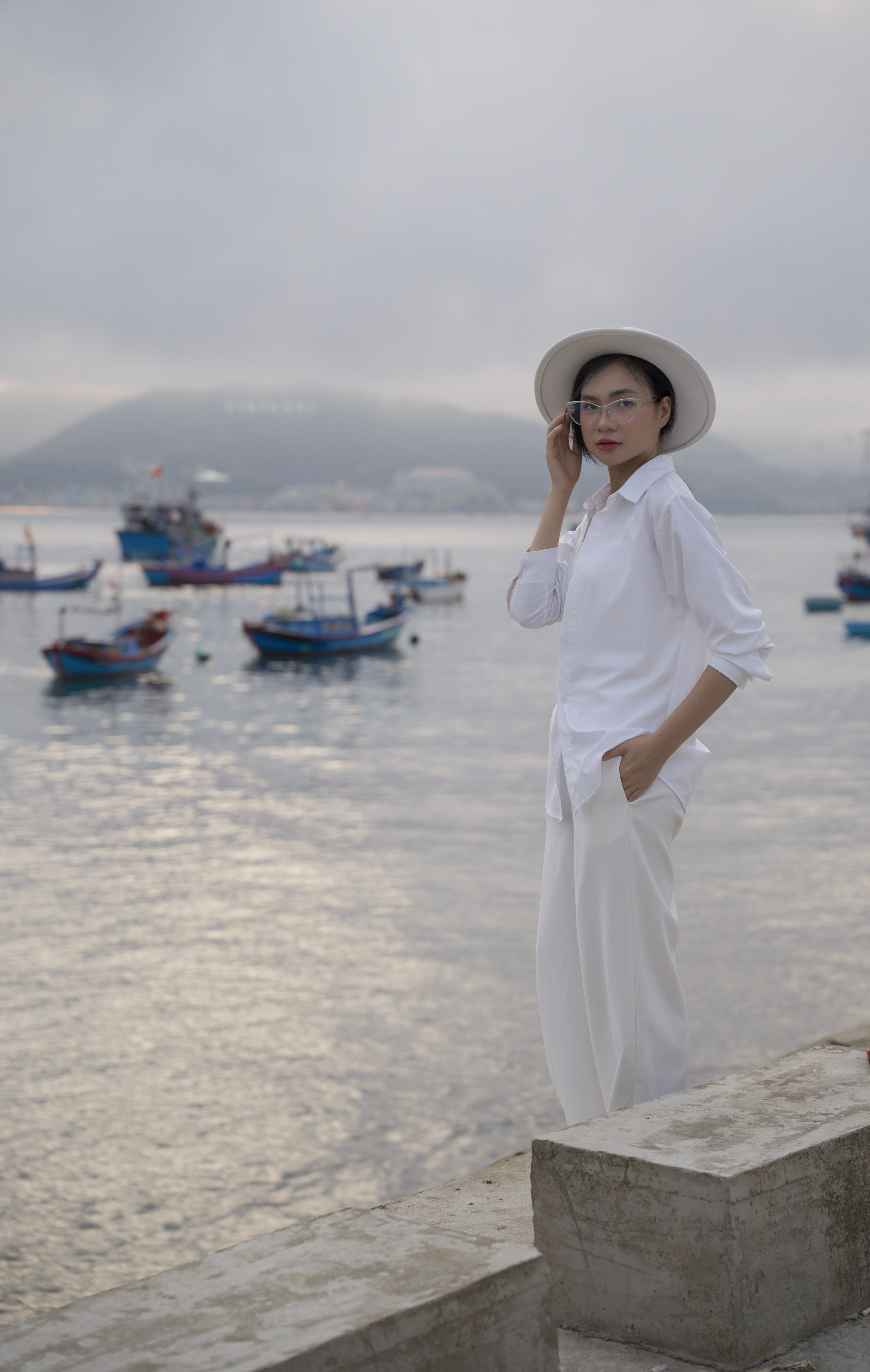 Woman in white outfit by the sea with boats in the background on an overcast day.