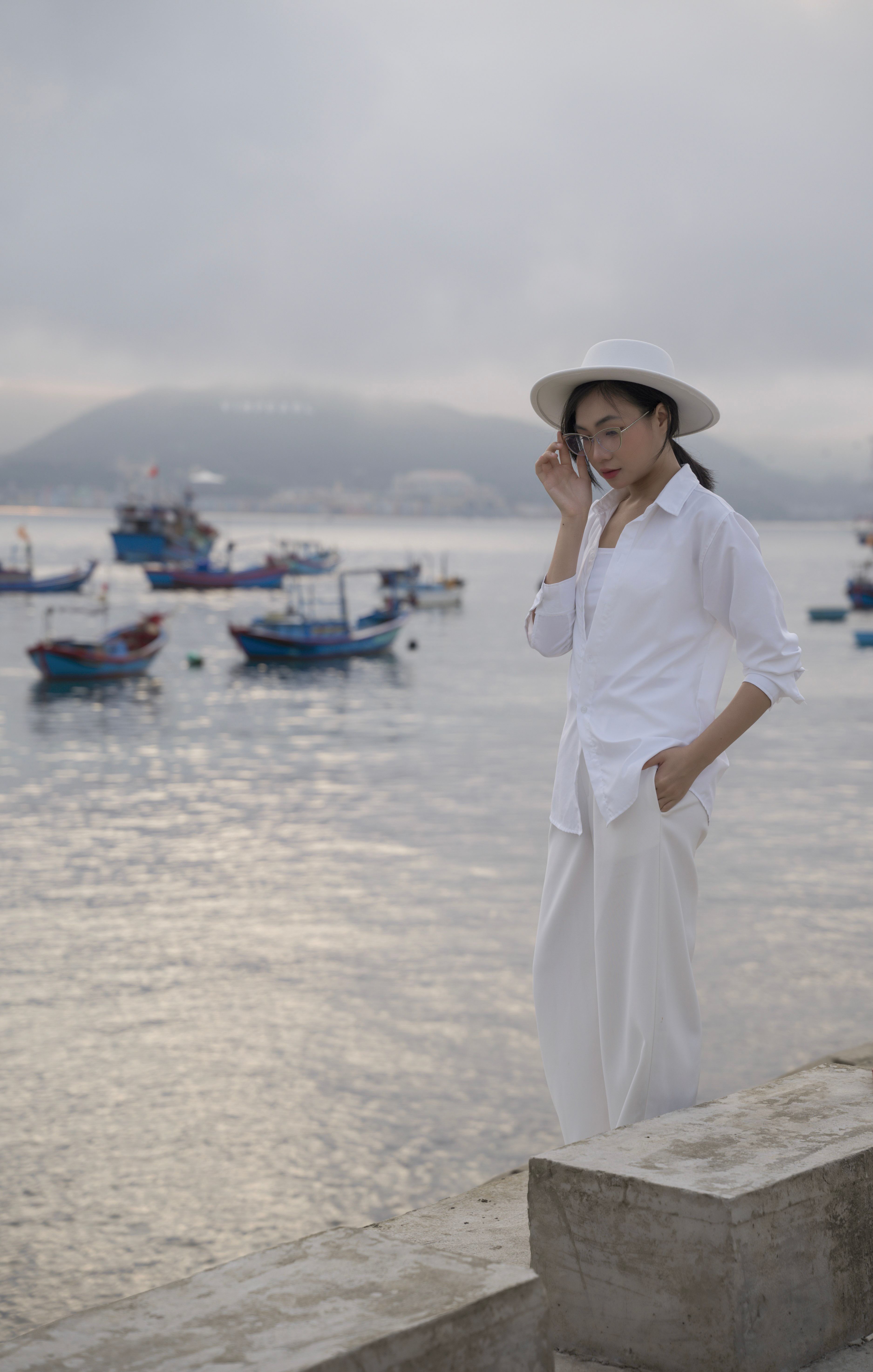 Woman in white clothing standing by a misty seaside with boats in the background.
