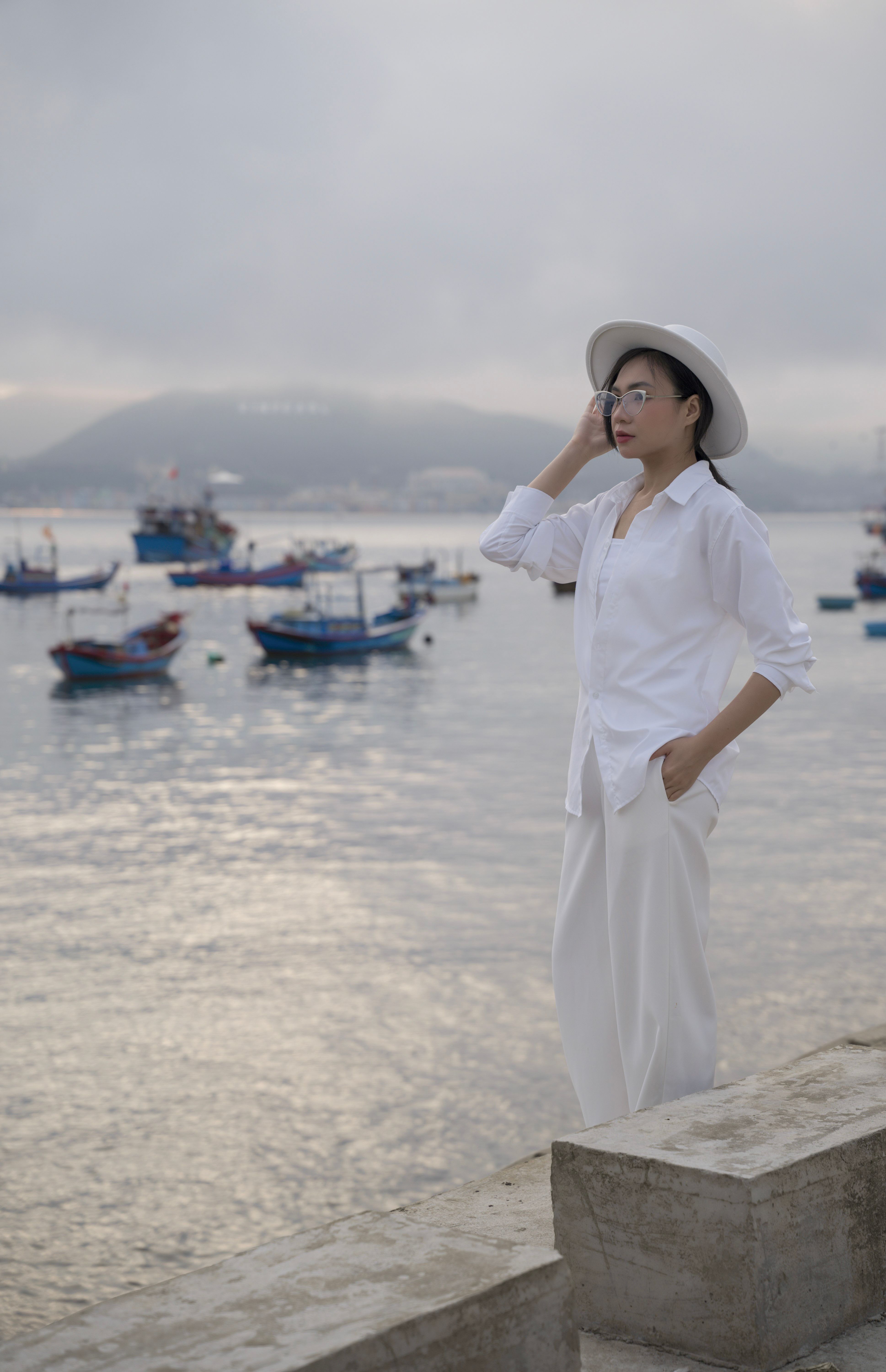Stylish woman in white outfit standing by the sea, surrounded by fishing boats at dusk.