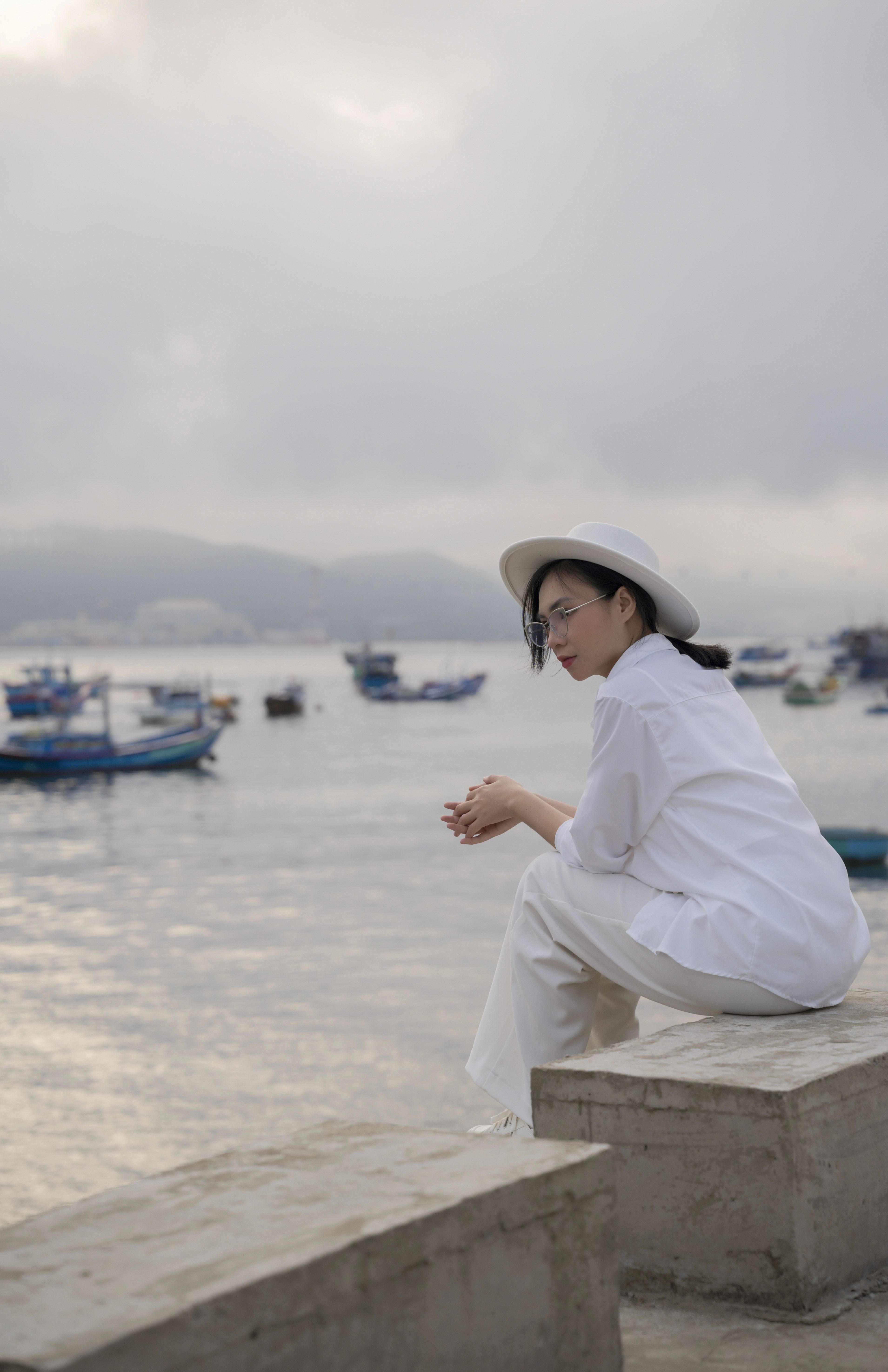 Woman in white clothing sitting by a misty harbor, contemplative mood.