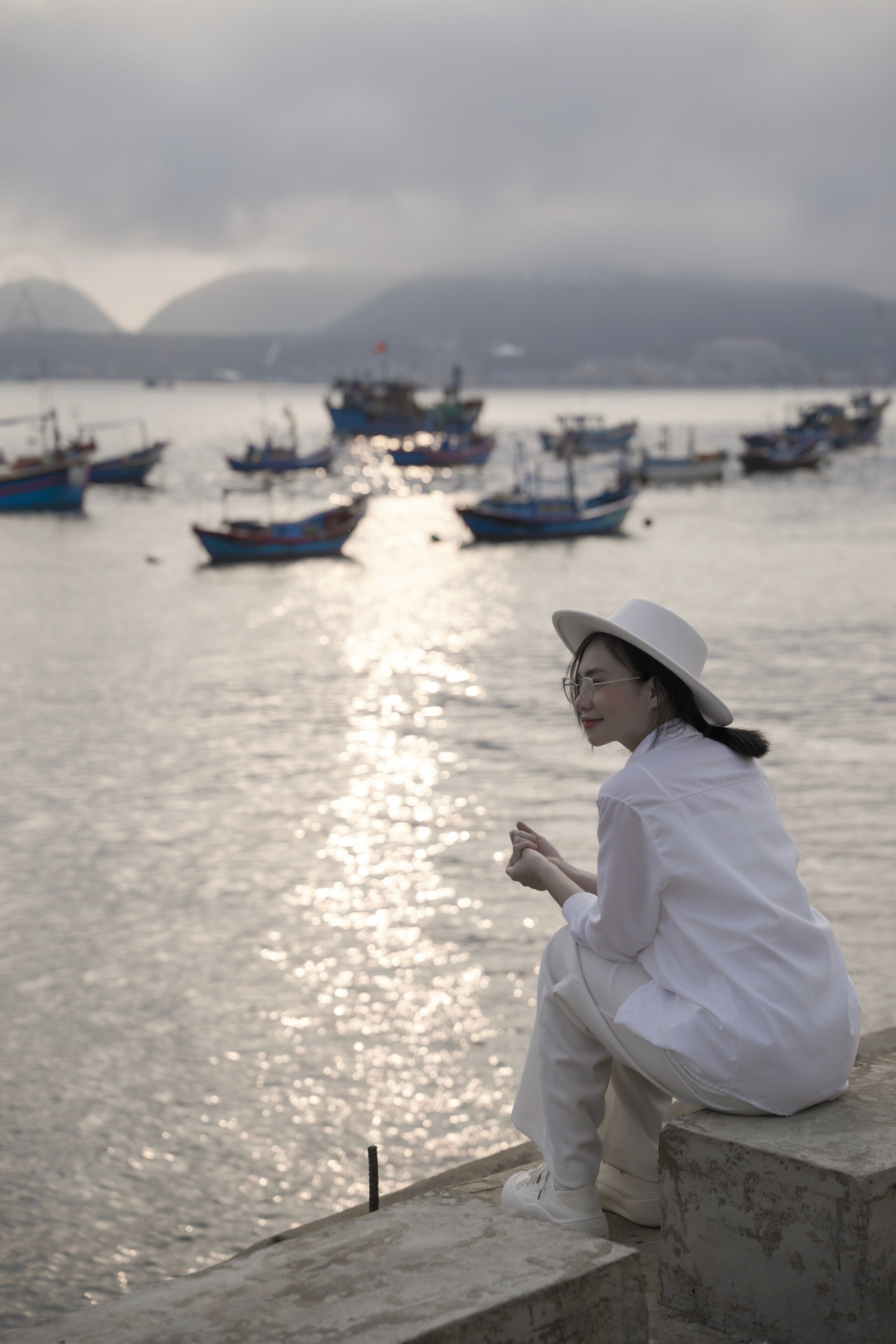 Woman in white sitting by sea, observing boats under cloudy sky.
