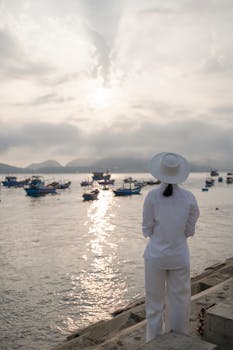 A woman in white gazing at boats on a tranquil harbor at sunset.