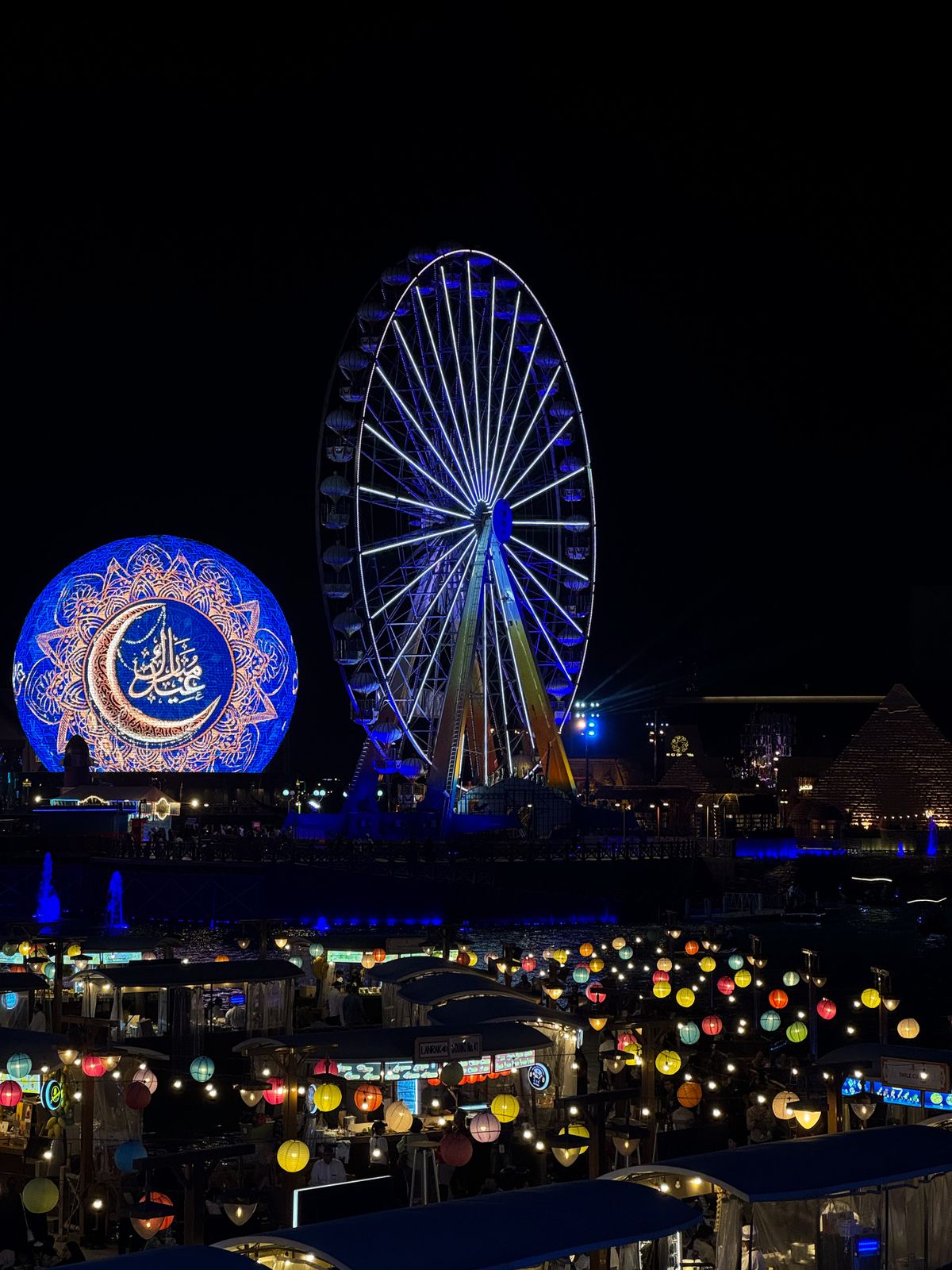 The Festival Wheel and the illuminated Global Village globe above lantern-lit stalls by the water at night