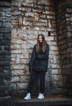 A young woman stands in winter clothing, smiling against a rustic stone wall, showcasing urban style.