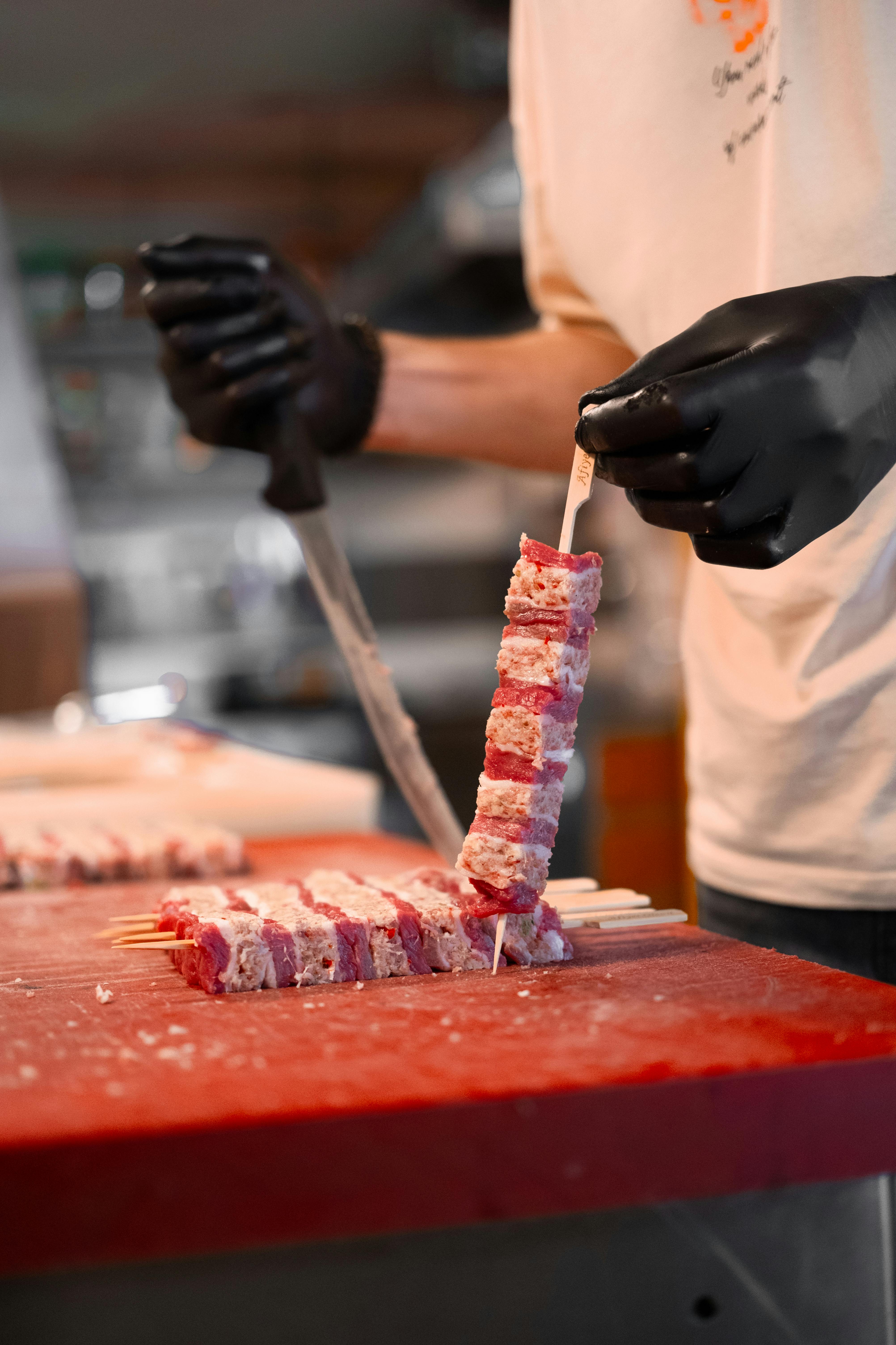 Chef skillfully cutting skewered red meat with a large knife, ready for grilling.