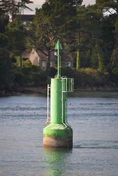 A solitary green buoy stands in calm coastal waters, surrounded by lush greenery and distant houses.