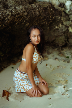 Young woman in patterned swimwear kneeling on a sandy beach by rocky cliffs.