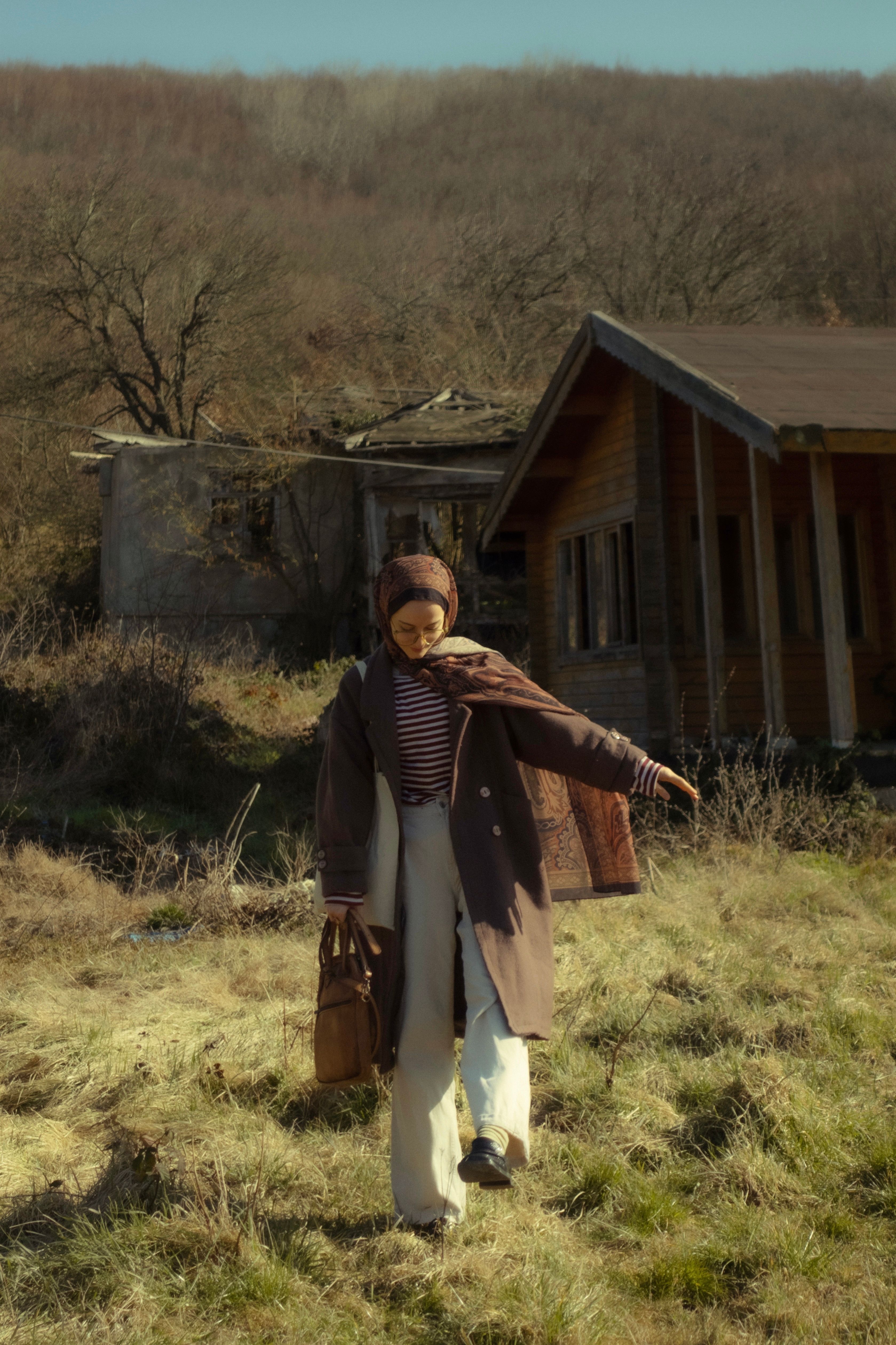A woman walks through an autumn field beside a rustic cabin, wearing warm clothing. Calm and peaceful scene.