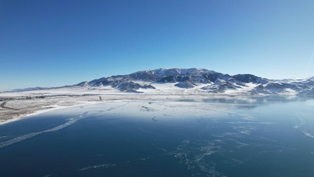 A breathtaking aerial view of snow-covered mountains and a serene lake under a clear blue sky.