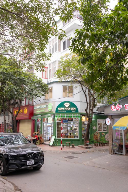 Free Explore a bustling Hanoi street lined with local shops, trees, and parked cars, capturing the essence of urban life. Stock Photo