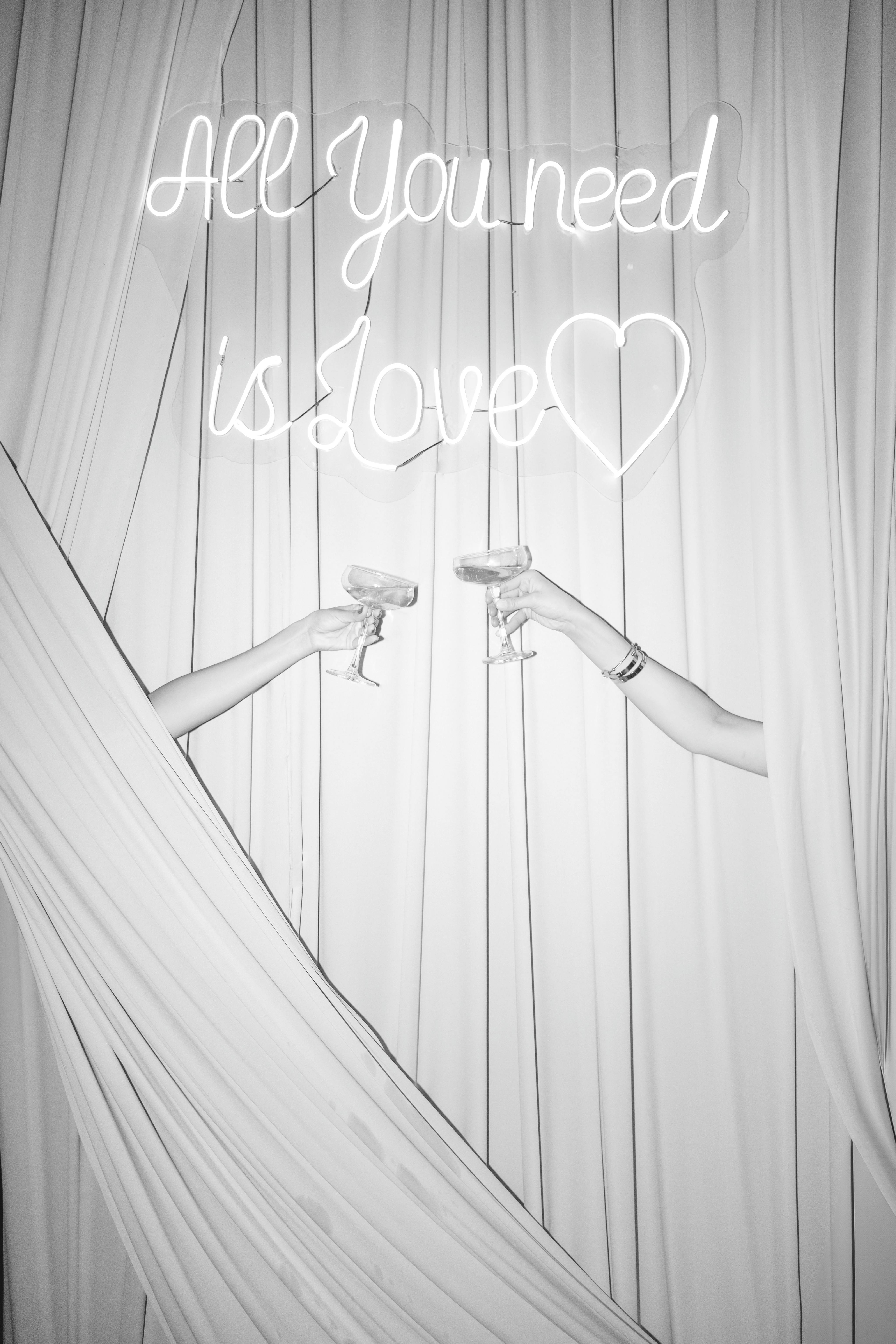 Two people toasting with champagne under 'All You need is love' neon sign.