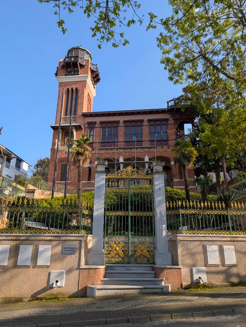 Free View of a historic red brick building with a tall tower amidst urban greenery on a sunny day. Stock Photo