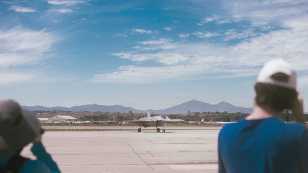 A fighter jet taxis on a runway with observers in the foreground and mountains in the background.