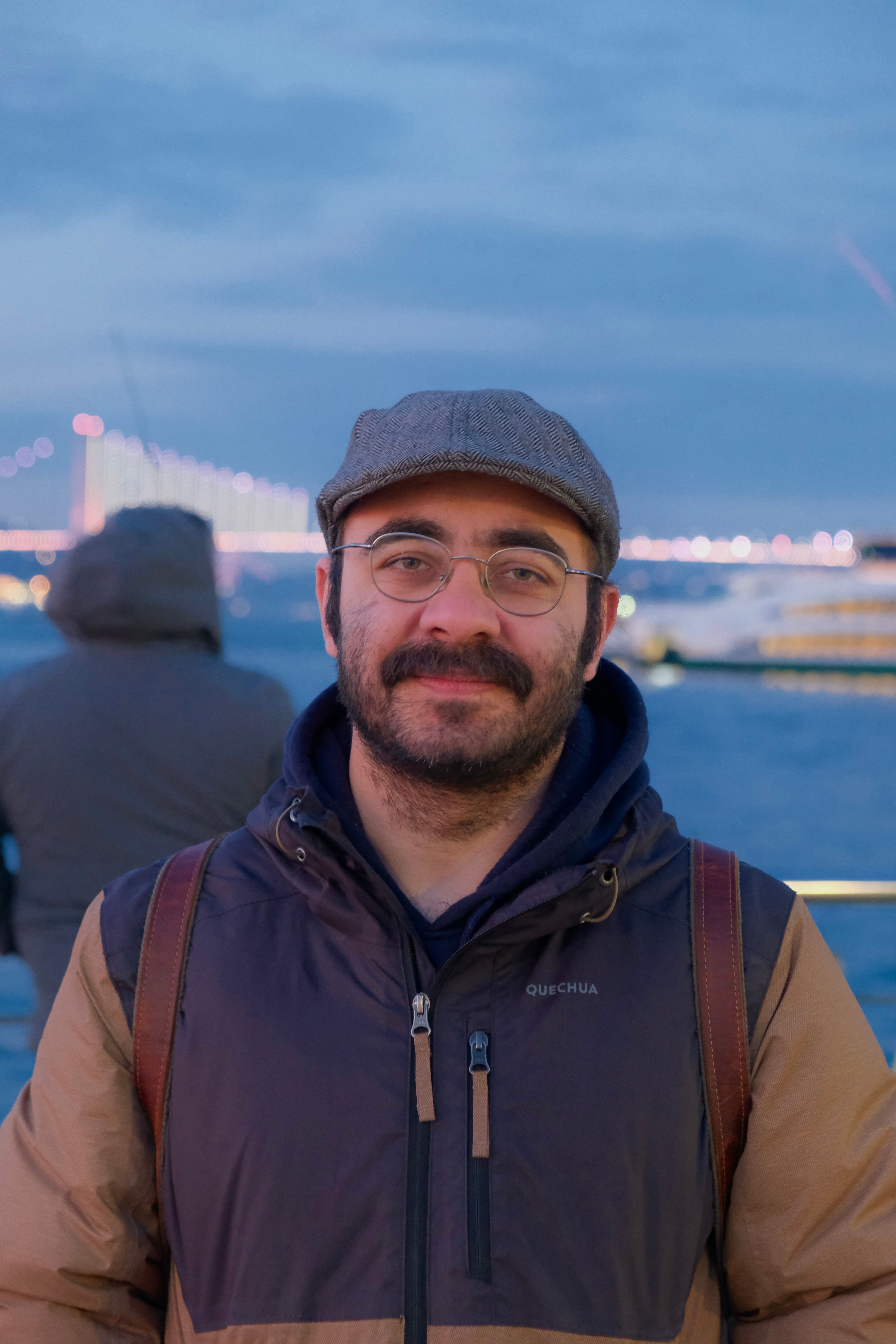 A bearded man smiles by the Bosphorus Bridge during twilight, capturing a serene Istanbul view.