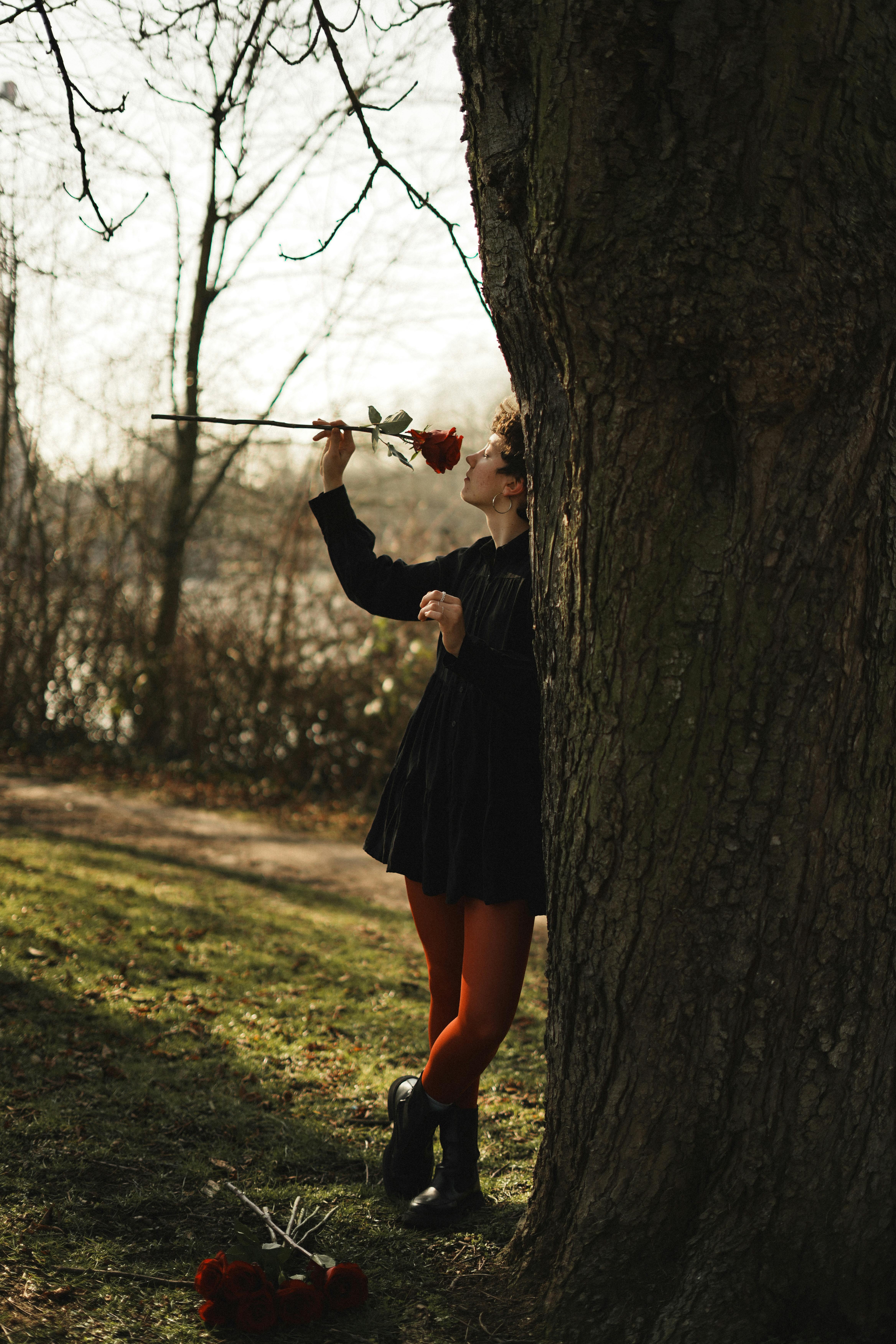 A person in a dark outfit enjoys a red rose by a large tree outdoors.