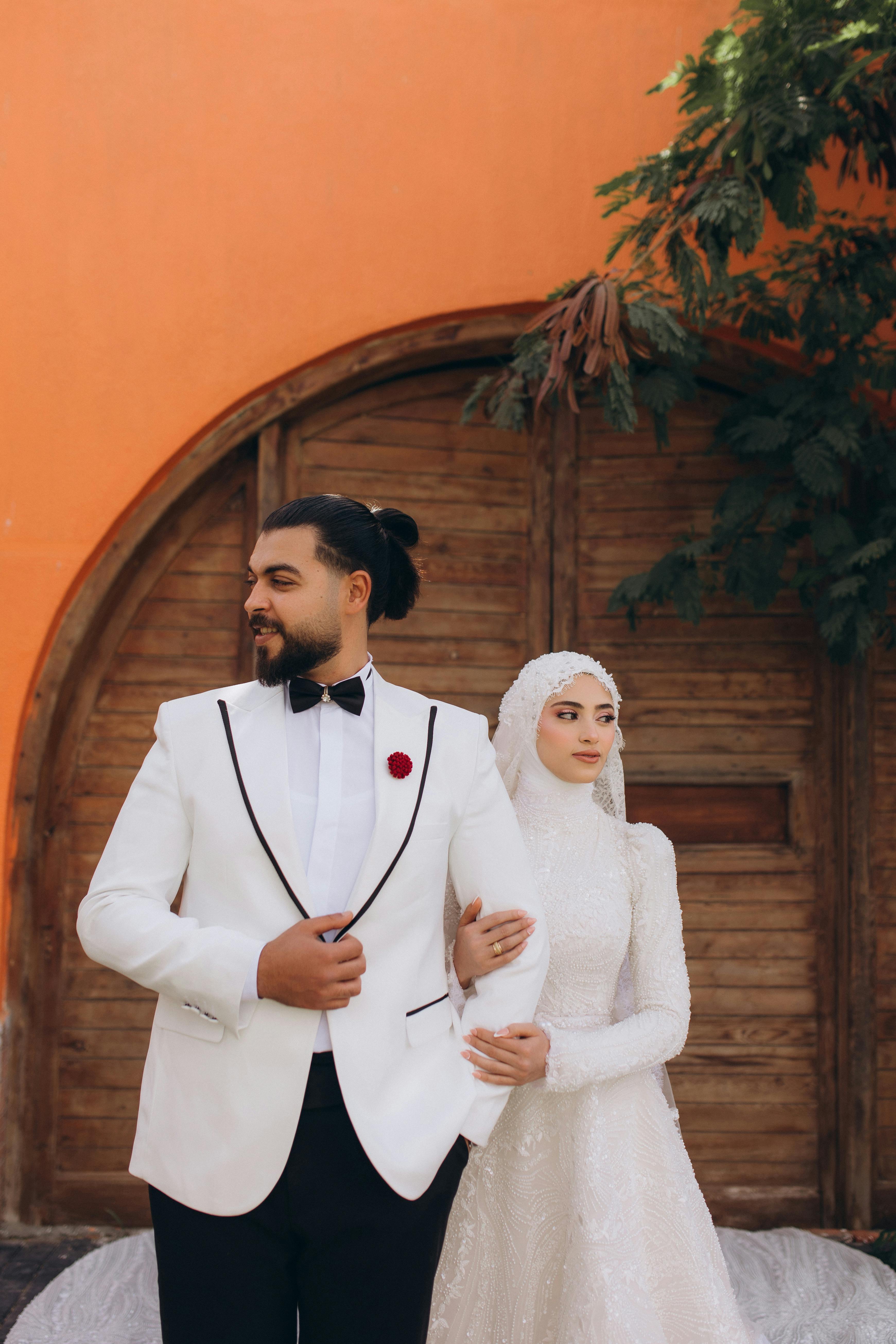 Elegant Muslim couple posing in formal wedding attire against a rustic wooden door and orange backdrop.