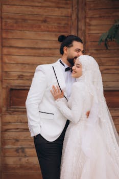Bride in hijab and groom in tuxedo embracing joyfully outdoors.