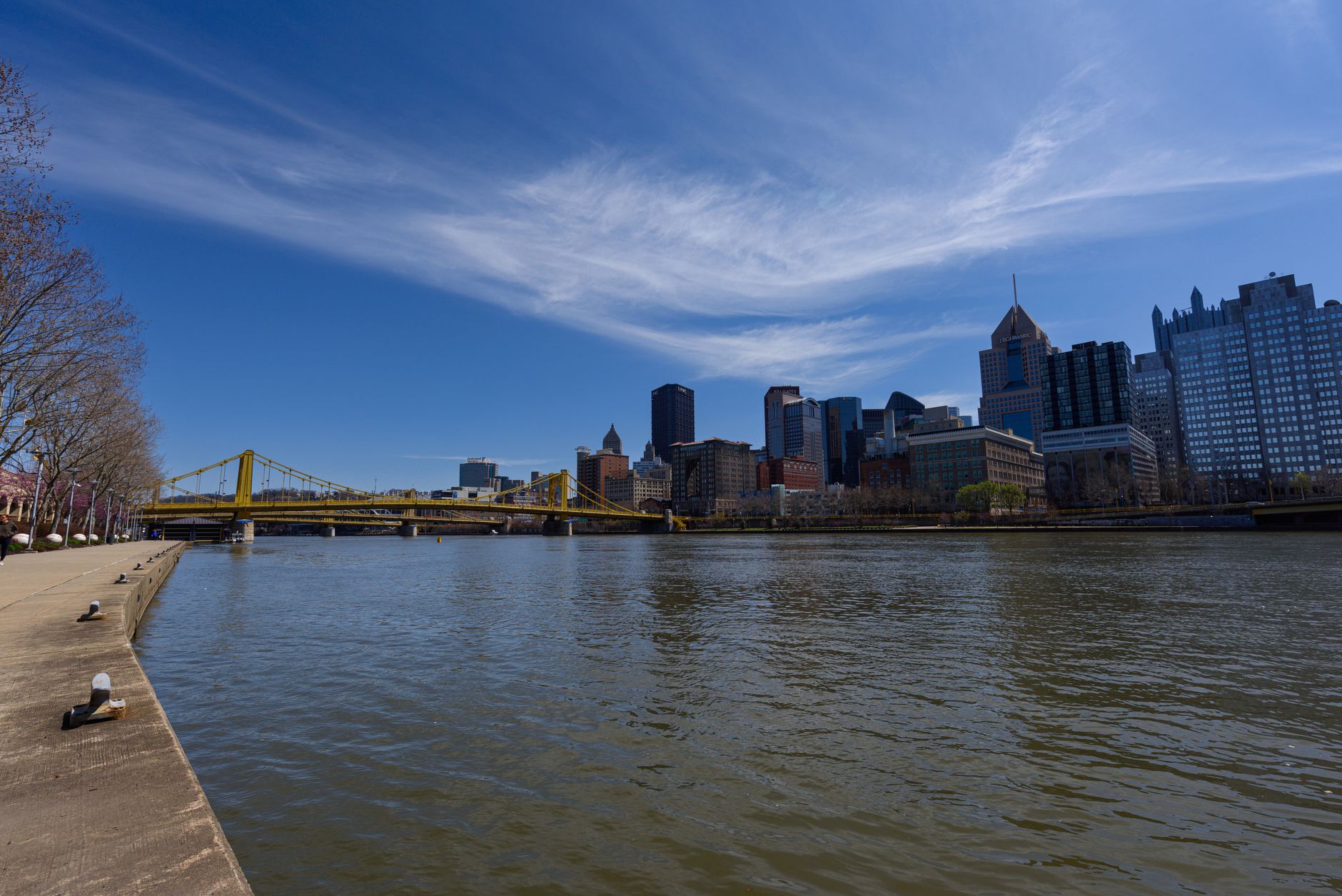 Scenic view of Pittsburgh skyline with yellow suspension bridge over calm river under clear blue sky.