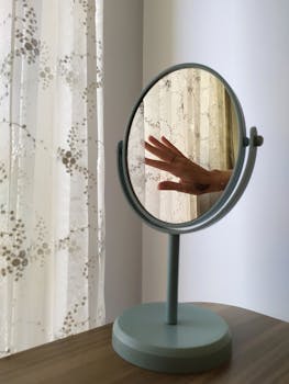 A hand reflected in a round mirror on a wooden table with light curtains in a minimalist room.