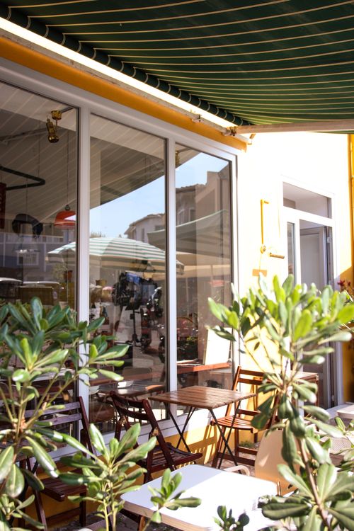 Free Sunlit café terrace with green awning, chairs, tables, and plants for a cozy outdoor ambiance. Stock Photo