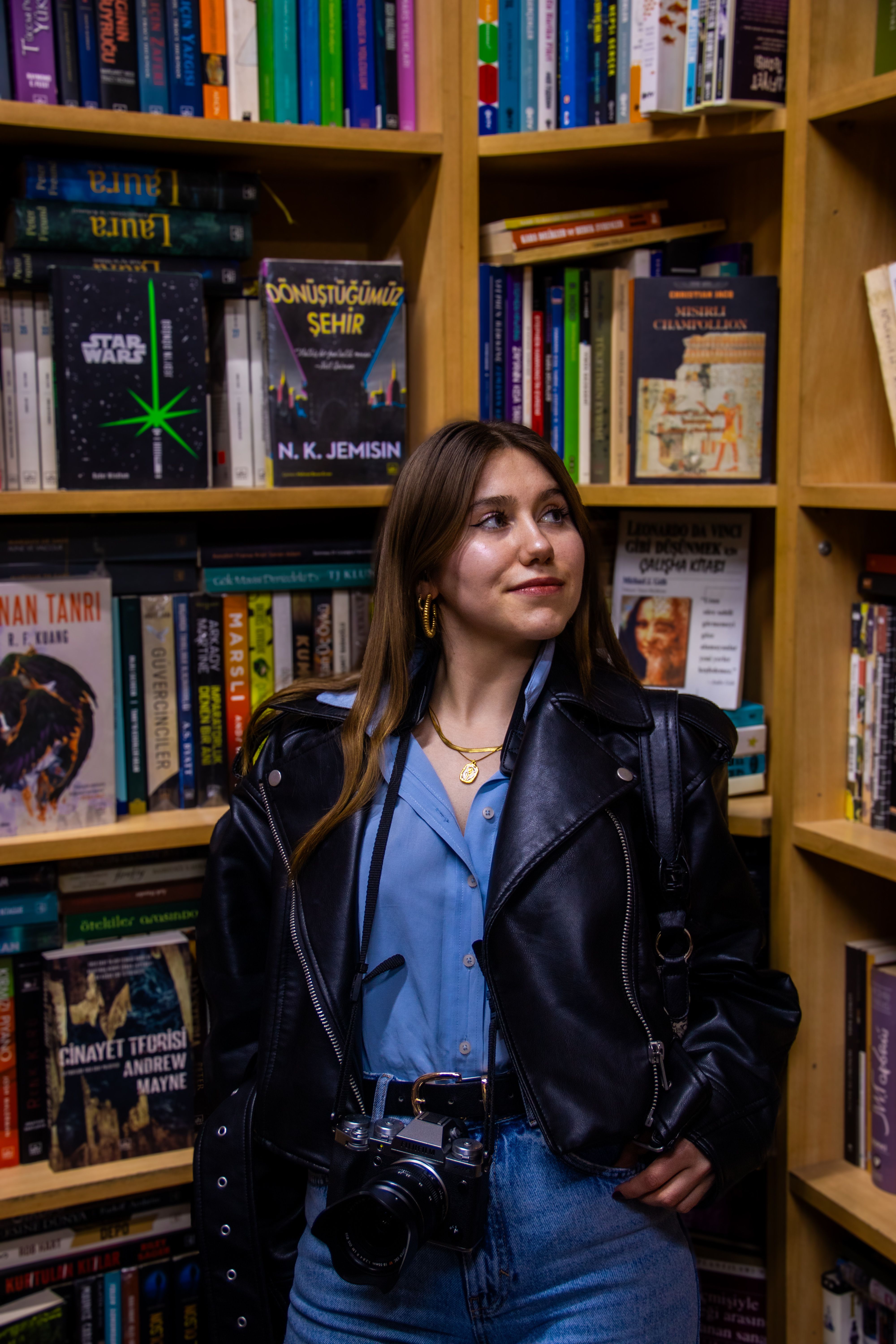Stylish woman in leather jacket poses with camera in a vibrant İstanbul bookstore among colorful bookshelves.
