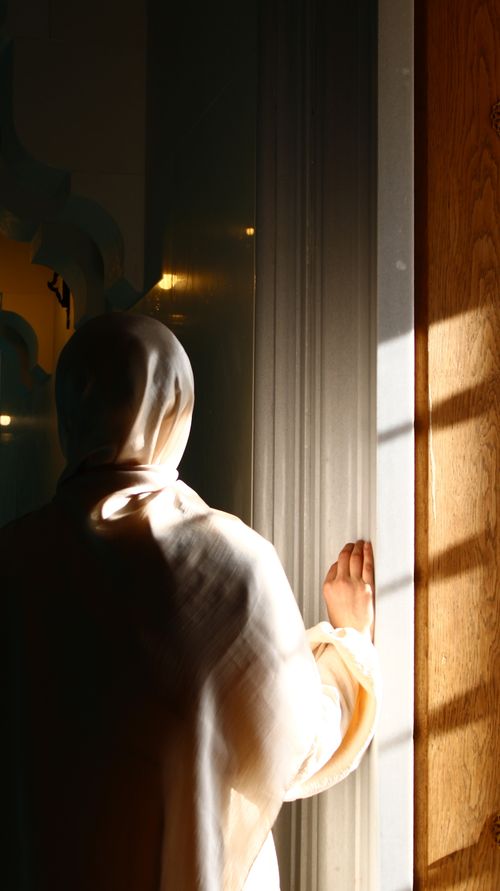 Free A woman in a hooded robe gazes out a sunlit window, casting a silhouette. Stock Photo