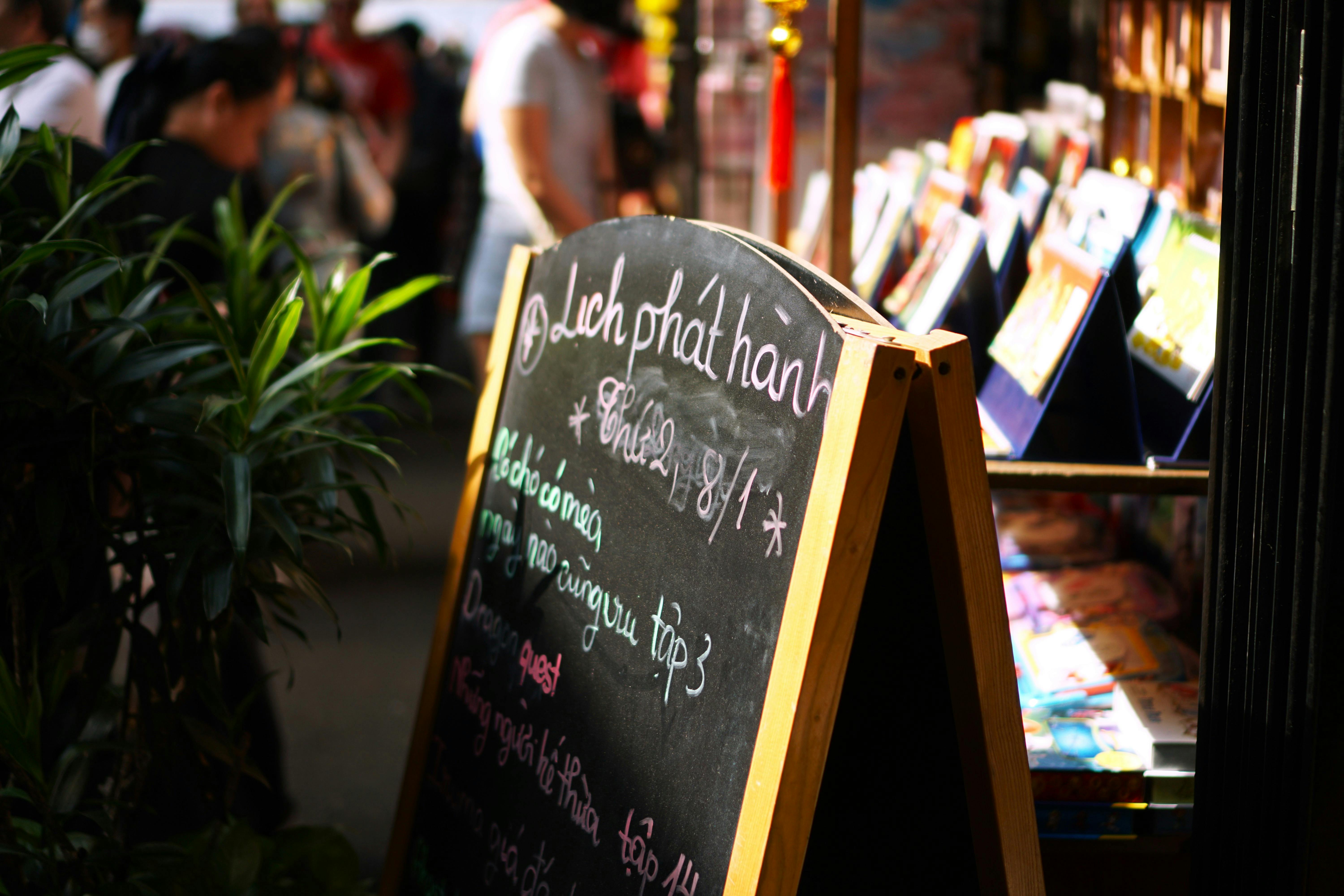 Chalkboard sign at an outdoor market with bustling background and plants.