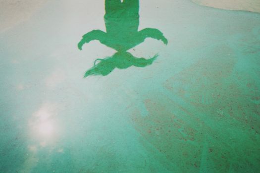 Artistic reflection of a person in a green-tinted water puddle, capturing an abstract silhouette.